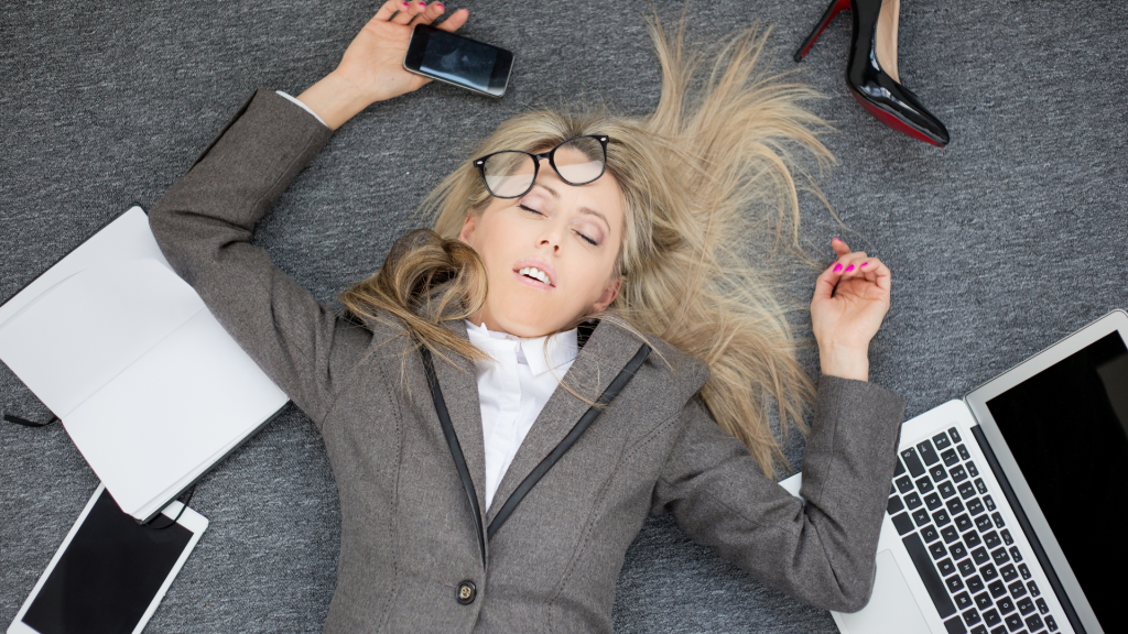This is a photo of a woman lying on the floor with her glasses hanging off, and devices strewn around. This image signifies burnout which is a characteristic of imposer syndrome. 
